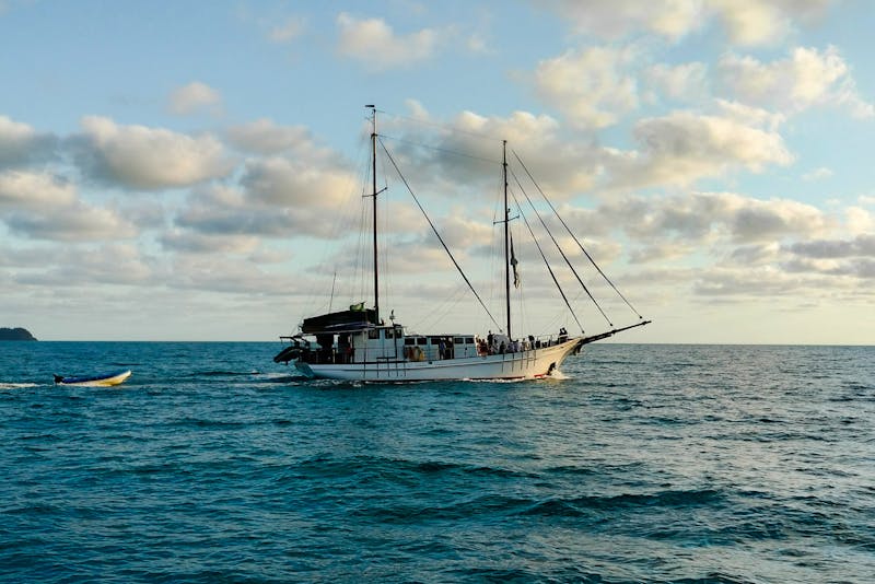 White sailing yacht cruising on calm blue sea at sunset with tender boat in tow.