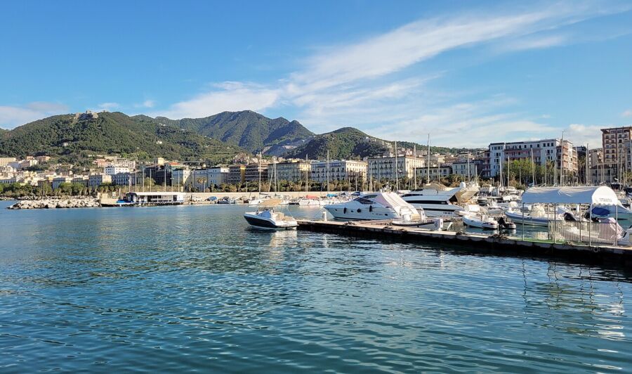Boats docked at Salerno Port with a city skyline and mountains in the background under a clear blue sky.