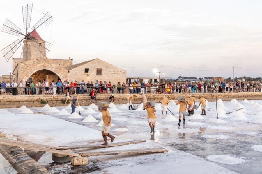 Workers harvest salt at a Salt pans of Trapani, surrounded by piles of salt under a cloudy sky. A crowd watches, and a traditional windmill stands nearby.
