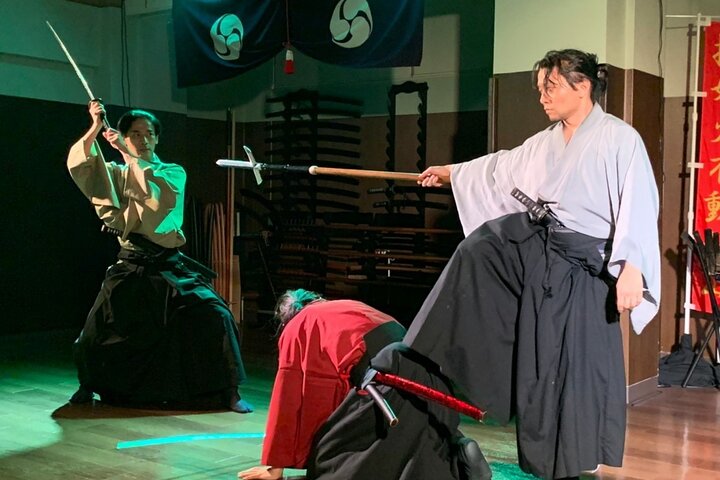 Three people in traditional Japanese attire perform a martial arts demonstration in a dojo. One holds a sword aloft, another a polearm, and one kneels.