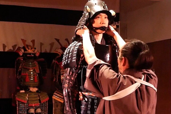 Person is being fitted with traditional samurai armor by another individual. The scene is serious, highlighting cultural heritage and craftsmanship.
