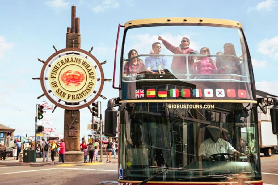 Tourists ride a double-decker bus near the Fisherman's Wharf sign in San Francisco. The scene is lively, with people enjoying the sunny day.