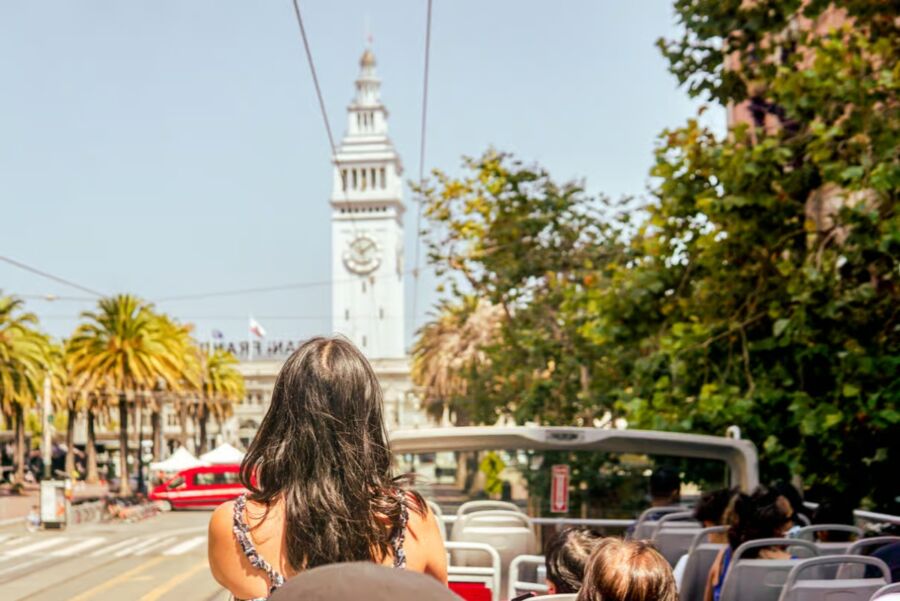 Woman with long dark hair rides an open-top bus, approaching a clock tower surrounded by palm trees. It's a sunny day, conveying a sense of exploration and leisure.
