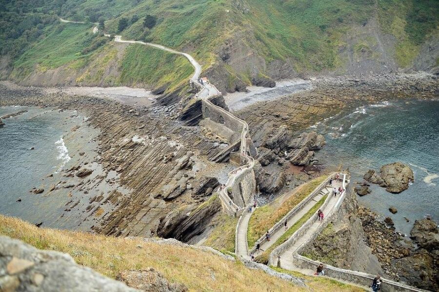 San Juan de Gaztelugatxe, Bay of Biscay, Basque coast of Spain narrow stone walkway winds along rugged cliffs, leading to a scenic hill with grassy vegetation. People walk along, enjoying the dramatic coastal views.