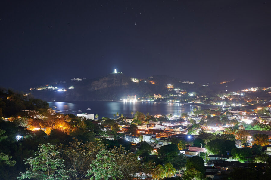 San Juan Del Sur at night, with sparkling lights reflecting on the water and a lively beach scene filled with energy 