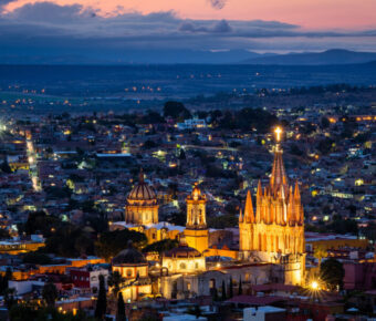 Aerial view of San Miguel de Allende at night, showcasing illuminated buildings and vibrant city lights against a dark sky.