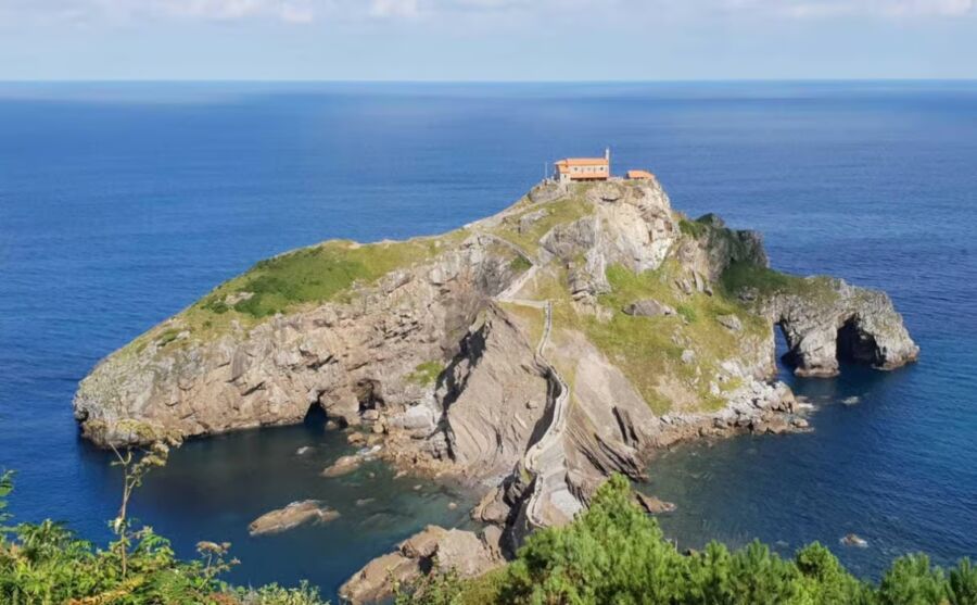 Rocky island with a winding stone staircase leads to a small orange-roofed building on top of the Sanctuary of San Juan de Gaztelugatxe, surrounded by serene blue ocean and sky.
