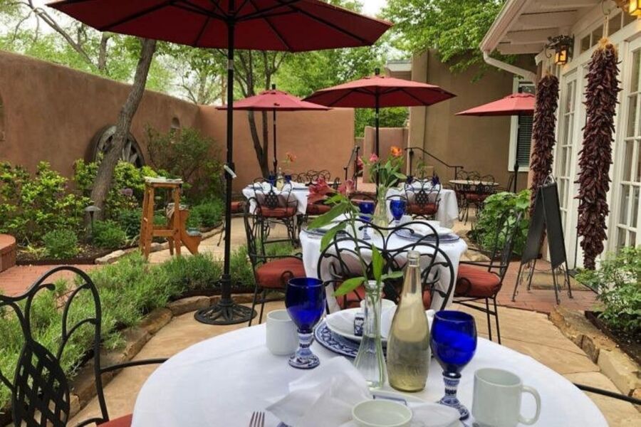 Gourmet Brunch in Charming Santa Fe restaurant patio with elegant table settings, featuring white tablecloths and blue glasses under red umbrellas. Lush greenery and a warm, inviting ambiance.