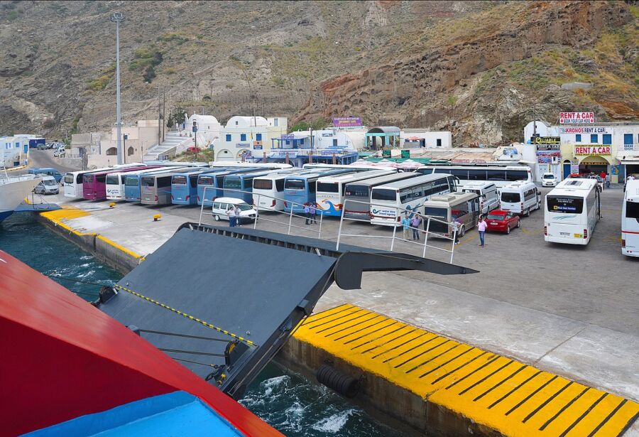 Santorini bus terminal with buses parked, travelers waiting, and a backdrop of traditional white buildings and blue skies