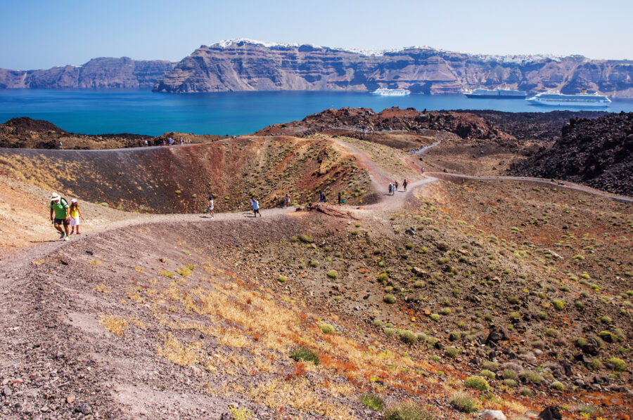 Hikers exploring the volcanic landscape of Santorini, Greece, with dramatic cliffs and the Aegean Sea in the distance