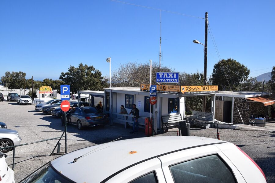 White car is parked at the Santorini Taxi Station, surrounded by other vehicles in the parking lot.