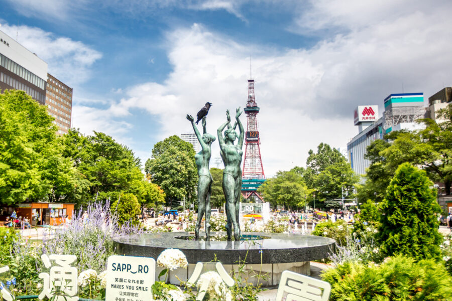 Sapporo Odori Park fountain with bronze statue, flowers, trees, and Sapporo TV Tower cityscape.