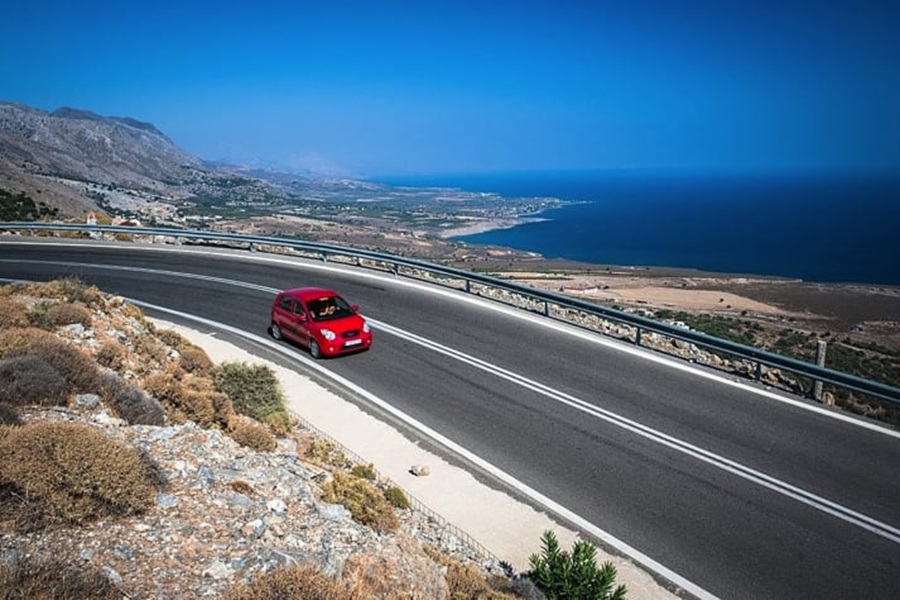 Coastal Mediterranean road trip with red car, blue sky, sea view, and mountainous backdrop.