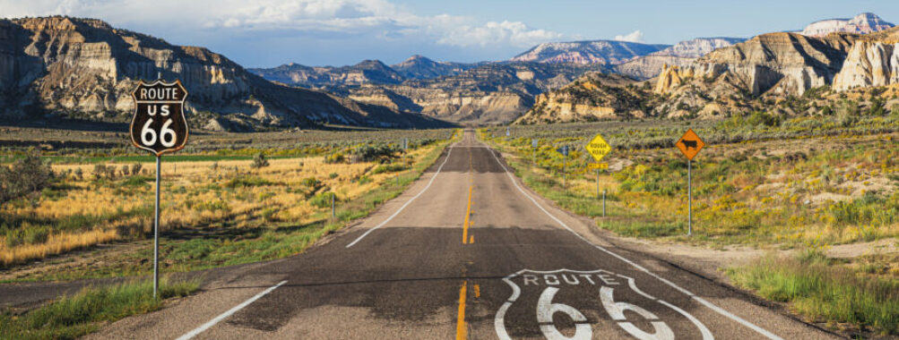 Straight desert road with "Route 66" painted on it, leading towards distant rocky mountains under a clear blue sky. Road sign reads "Route US 66."