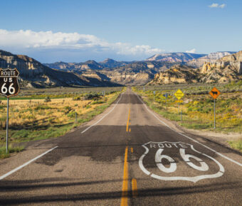 Straight desert road with "Route 66" painted on it, leading towards distant rocky mountains under a clear blue sky. Road sign reads "Route US 66."