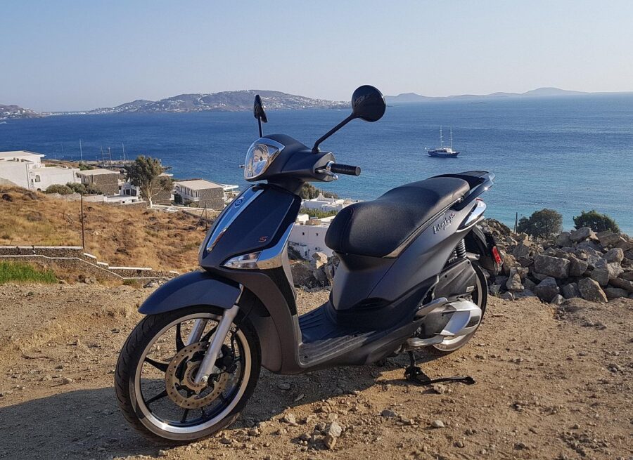 Modern Vespa scooter on a rocky cliff, overlooking stunning Mykonos coastal landscape.