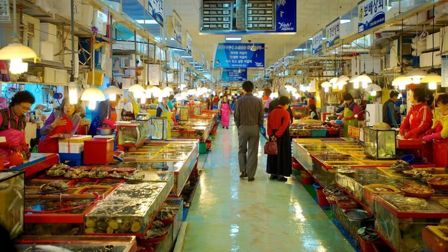 Busy indoor Korean seafood market with fresh fish stalls, shellfish tanks, vendors and shoppers.