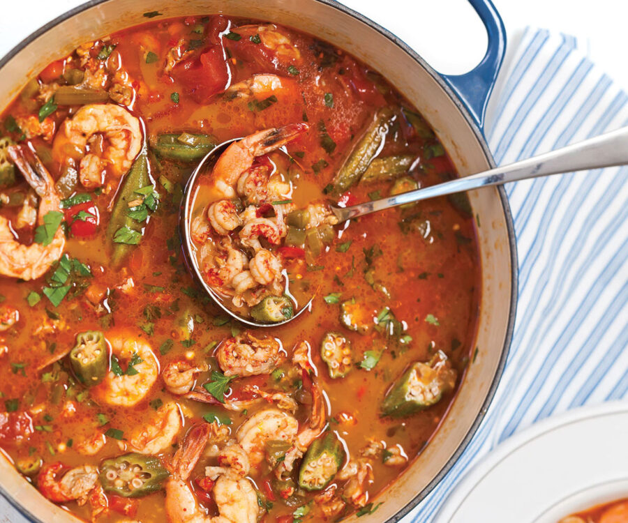 Seafood stew with shrimp, okra, peppers, and herbs in a pot on a striped cloth.