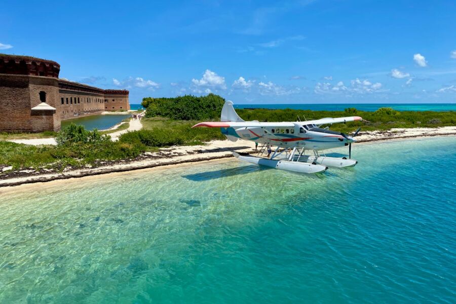 Seaplane docked by tropical beach with historic fort, lush greenery, and turquoise water.