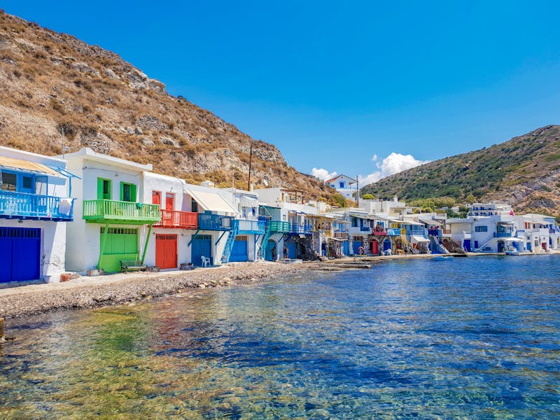 Whitewashed Mediterranean harbor houses with colorful balconies along turquoise pebble shoreline.