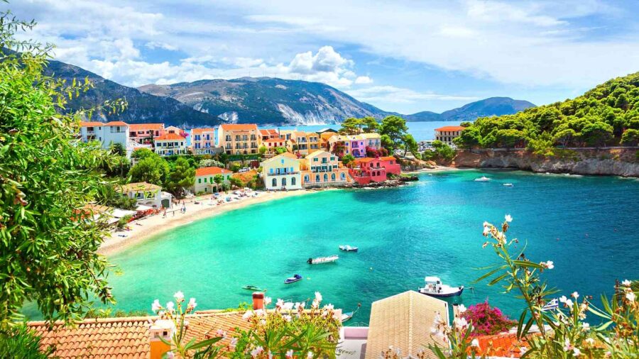 Mediterranean coastal town with colorful buildings, sandy beach, turquoise waters, and lush hills.