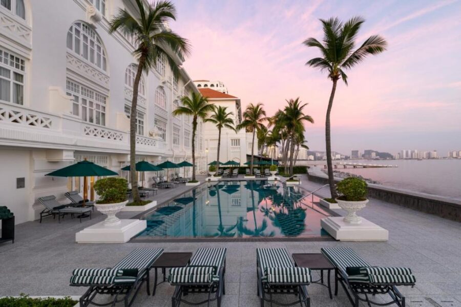 Luxury colonial-style seaside hotel infinity pool at sunset with palm trees and city skyline views.