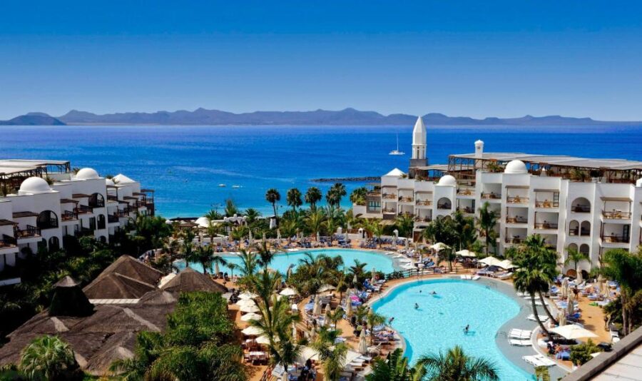 Luxury beachfront resort with lagoon pool, palm trees, ocean view, and Mediterranean-style hotel buildings.