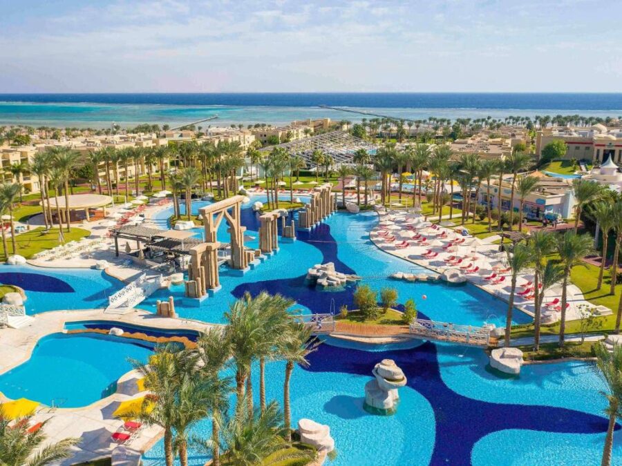 Luxury seaside resort with lagoon-style swimming pools, palm trees, sun loungers, and oceanfront views.