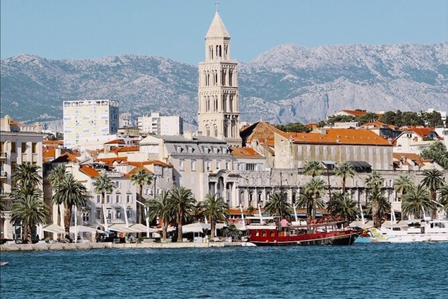 Mediterranean coastal town with palm-lined promenade, historic bell tower, boats, and mountain backdrop.
