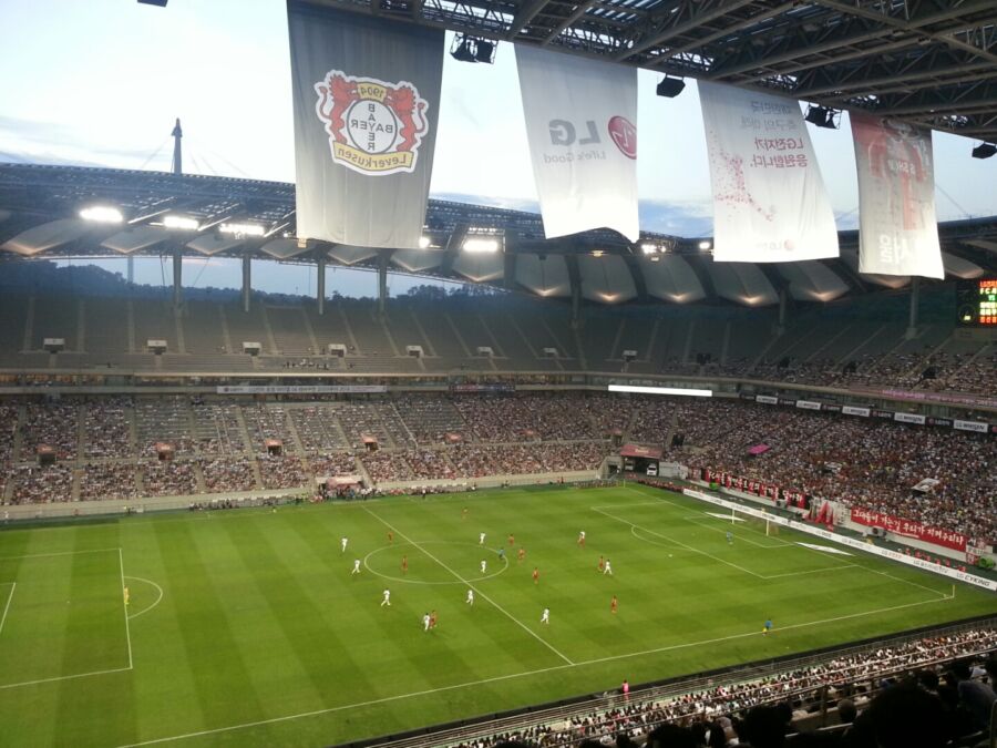 The Seoul Football Game stadium bustling with enthusiastic fans, banners waving, and players warming up on the field