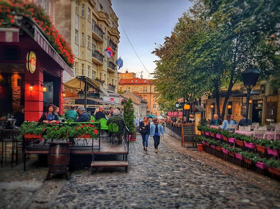 Charming cobblestone street in Skadarlija District, vibrant cafes, floral decor, lively evening ambiance.