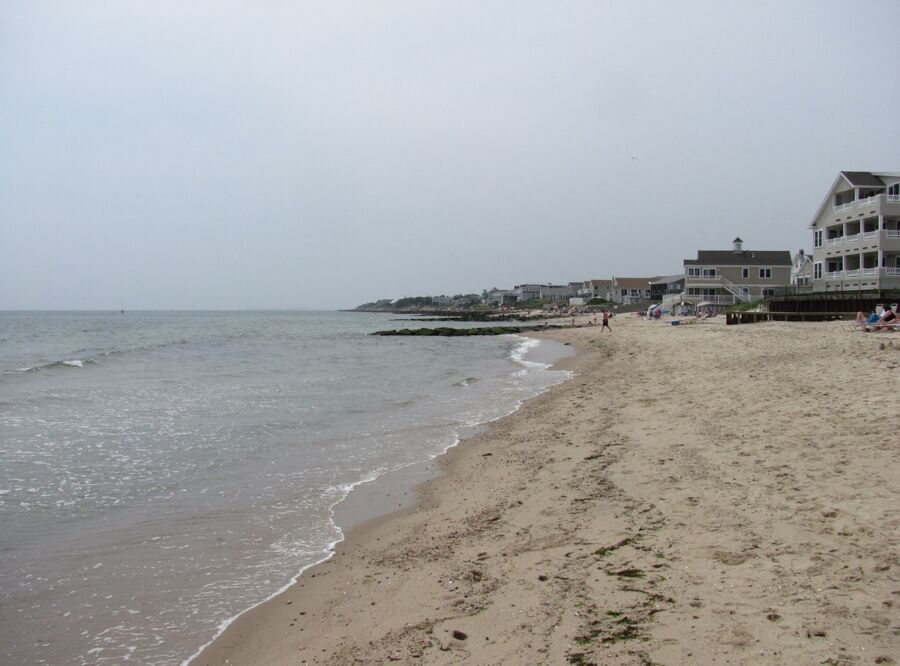 Tranquil sandy Dennis Port beach with gentle waves and seaside homes under overcast sky.