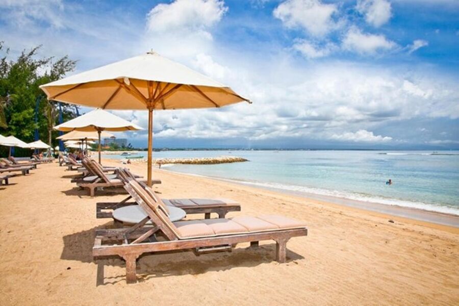 Golden sandy Sanur beach with lounge chairs, umbrellas, azure sea, and palm trees on a sunny day.