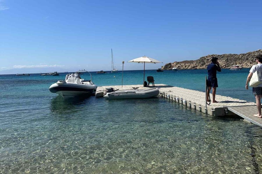 Seaside serenity: wooden dock, motorboats, turquoise waters, sunny sky, Mykonos beach scene.