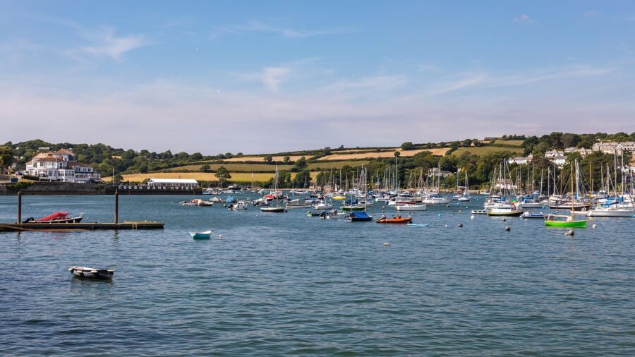 Bustling Falmouth harbor scene with boats, blue sky, green hills, and quaint coastal architecture.