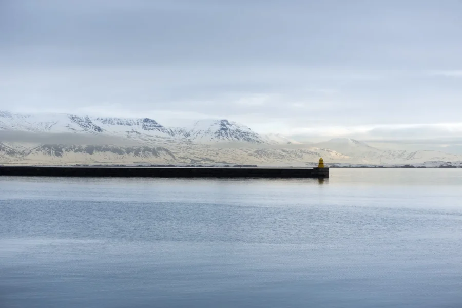 Serene coastal landscape with lighthouse, calm water, mountain range, and muted colors in Iceland.