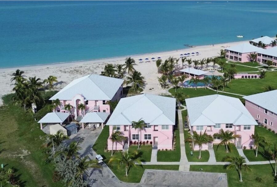Tropical beachside resort with pink buildings, white roofs, palm trees, pool, and ocean views.