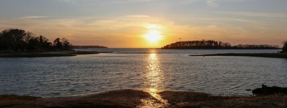 Cape Cod sunset over calm coastal landscape with island, reflecting golden hues on water.