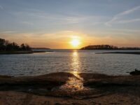 Cape Cod sunset over calm coastal landscape with island, reflecting golden hues on water.