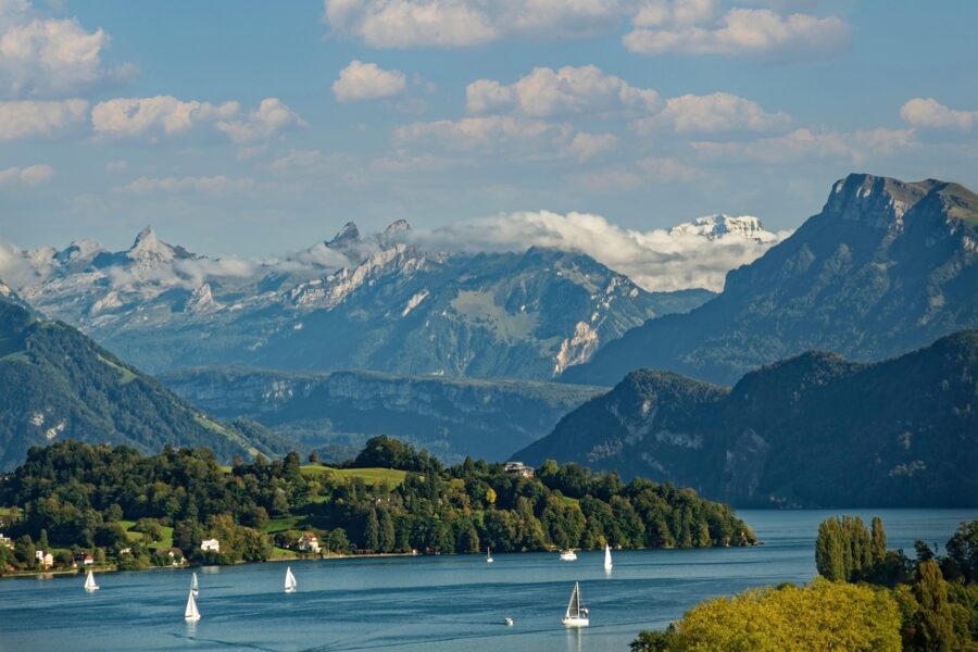 Serene Swiss lake with sailboats, lush greenery, rolling hills, and majestic mountain backdrop.