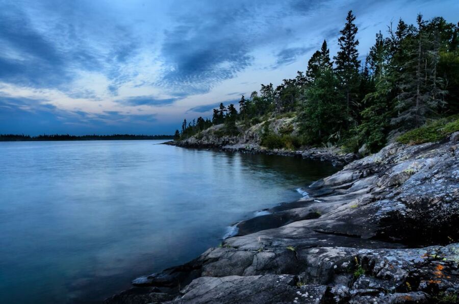 Twilight lakeside scene with calm water, rocks, trees, and colorful sky reflections.