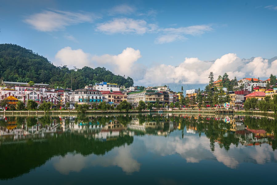 Tranquil emerald lake reflecting scenic hillside town, colorful buildings, forested mountains and blue sky.