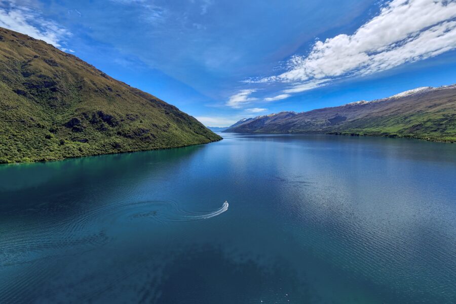 Aerial view of tranquil mountain lake with boat, forested slopes, snowcapped peaks and blue sky.