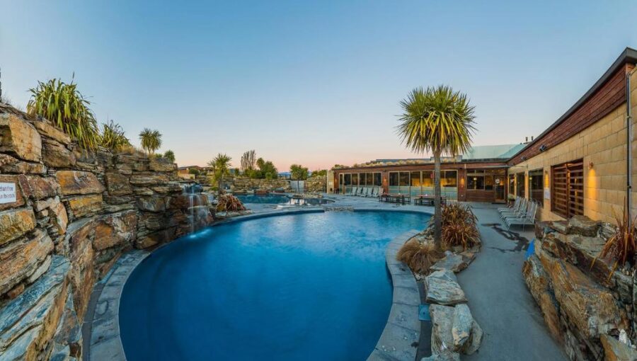 Luxury resort lagoon pool at dusk with waterfall, palm trees, lounge chairs and warm lighting.