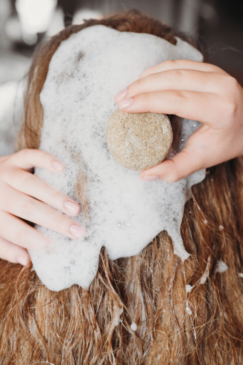 Person washing hair with a round shampoo bar. Soapy lather covers long, brown hair. Hands hold the bar, creating a clean and refreshing feel.