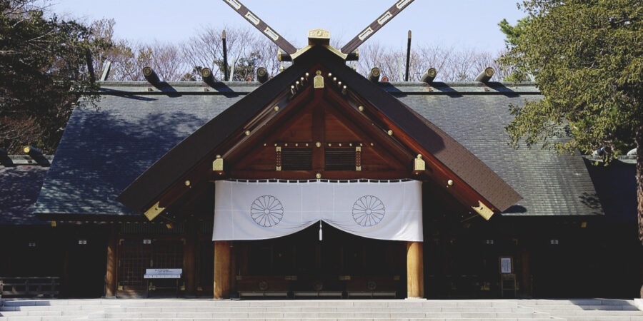 Front view of traditional Japanese Shinto shrine with dark wooden roof, white curtain, and trees.