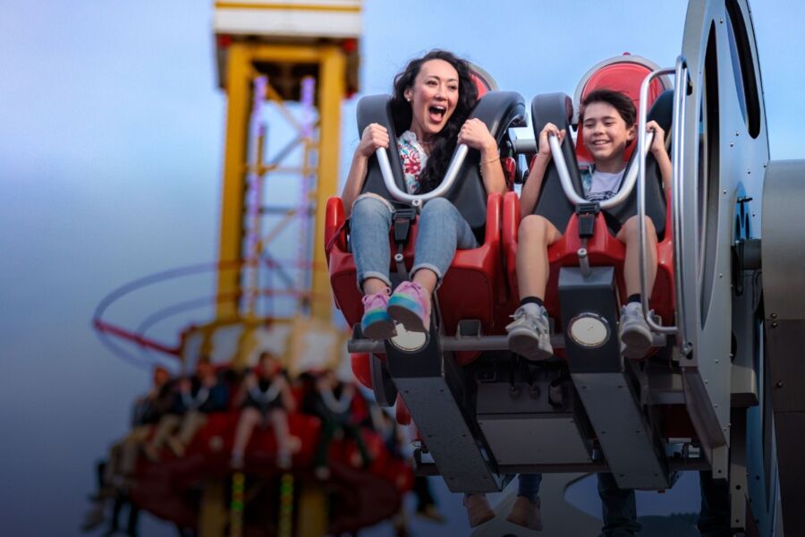 Woman and a child on Six Flags Fiesta Texas ride, smiling and excited. Background features blurred people on a spinning ride, sky is clear blue.