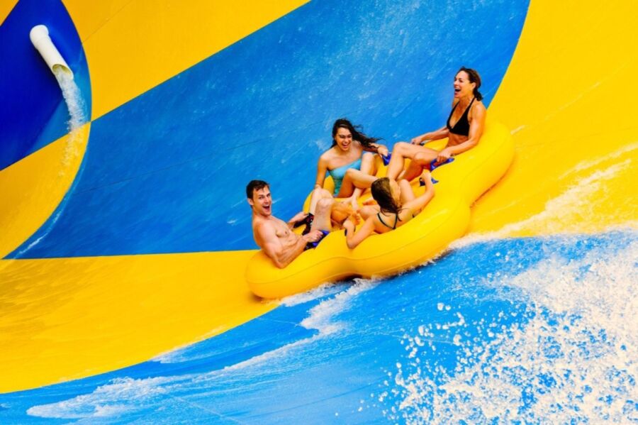 Four people excitedly ride a yellow raft down a vibrant blue and yellow water slide at Six Flags Fiesta Texas Waterpark. Water splashes around them, adding to the thrill and fun.