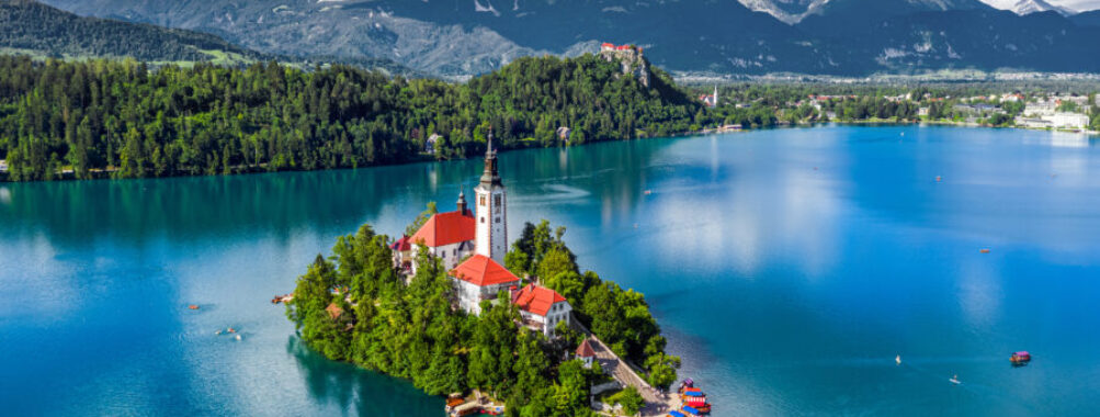 Aerial view of Lake Bled with the church, pletna boats, Bled Castle, and Julian Alps on a sunny summer day.