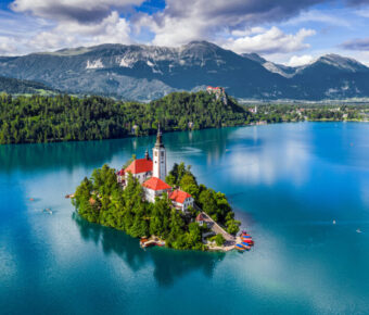 Aerial view of Lake Bled with the church, pletna boats, Bled Castle, and Julian Alps on a sunny summer day.